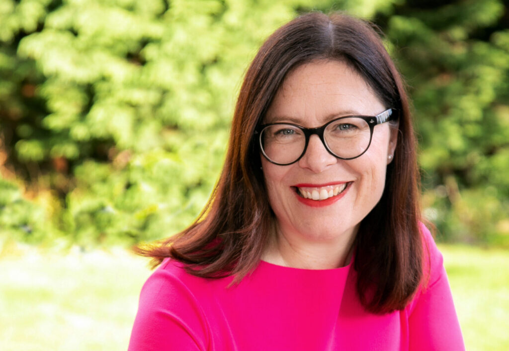 A woman with shoulder length brown hair wearing glasses and a pink top